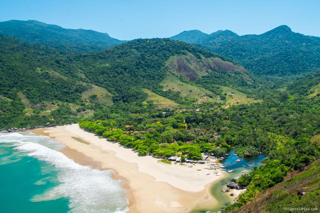 Vista aérea de uma praia tropical com colinas verdes, mar azul claro, areia branca e um pequeno rio desaguando no oceano. Essa é a Praia do Bonete, acessada por uma das trilhas em SP.