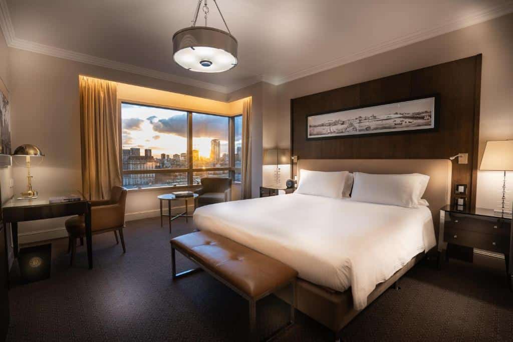 Quarto amplo com vista da cidade no Alvear Icon Hotel com cama de casal, mesinha e luminária de ambos os lados, poltrona com mesinha e janela grande, para repesentar hotéis perto da Casa Rosada em Buenos Aires.