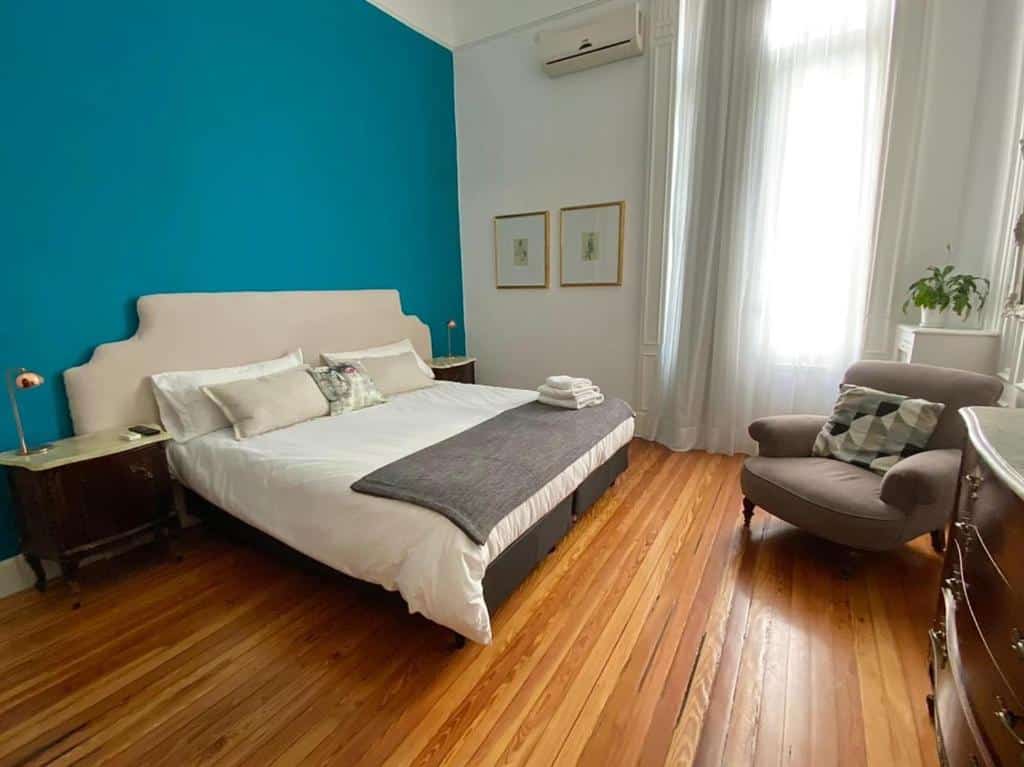 quarto do Hotel Chemin com cama de casal, mesinha e luminária de ambos os lados com chão de madeira, poltrona, quadros na parede e janela ampla com cortinas claras combinando com o verde da parede