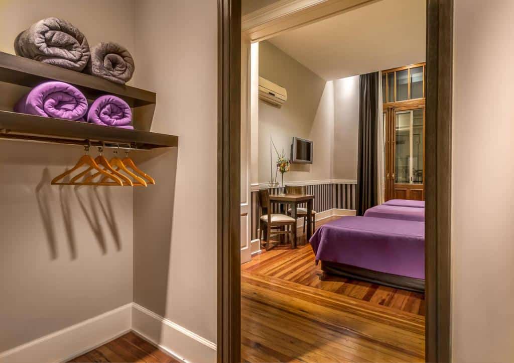 quarto do Up Tribeca visto a partir do closet com decoração violeta, cama de casal, janela grande acima e mesa com cadeiras de madeira em um ambiente amplo