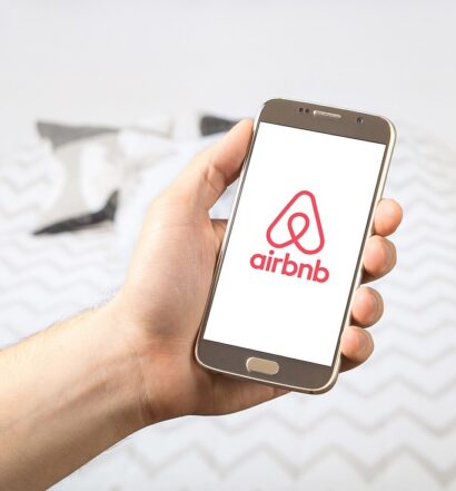 Quarto com uma cama de casal, atrás de uma mão segurando um celular com o logo do Airbnb. Para ilustrar o post se o Airbnb é confiável
