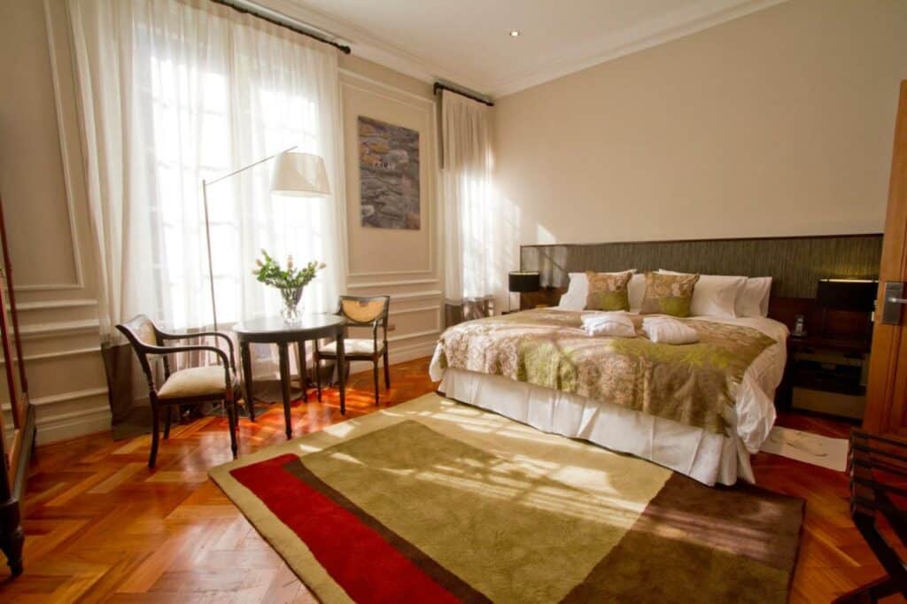 Quarto do Casa Bueras Boutique Hotel com cama no centro do quarto com uma cômoda em cada lado com luminária e a frente da cama do lado esquerdo uma mesa redonda com duas cadeiras.