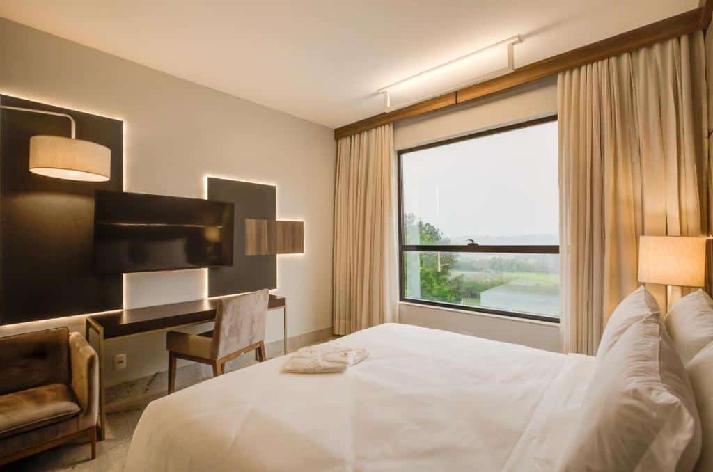 Quarto do DoubleTree by Hilton com um cama de casal e de frente um painel com tv, uma mesa com uma cadeira e uma poltrona. Já do lado direito da cama tem uma janela de vidro com cortinas abertas.