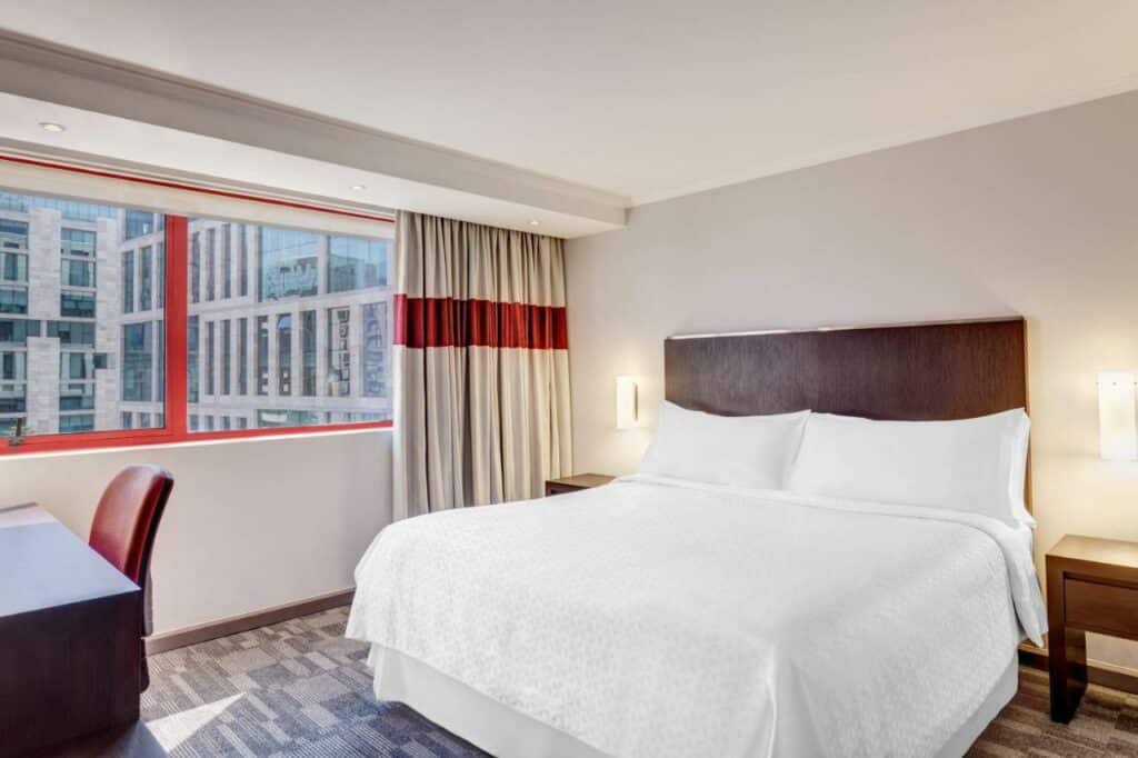 Quarto do Four Points by Sheraton Santiago com cama de casal do lado direito da imagem com uma cômoda em cada lado da cama e em frente a cama do lado direito uma mesa de trabalho com cadeira. Representa hotéis 4 estrelas em Santiago.