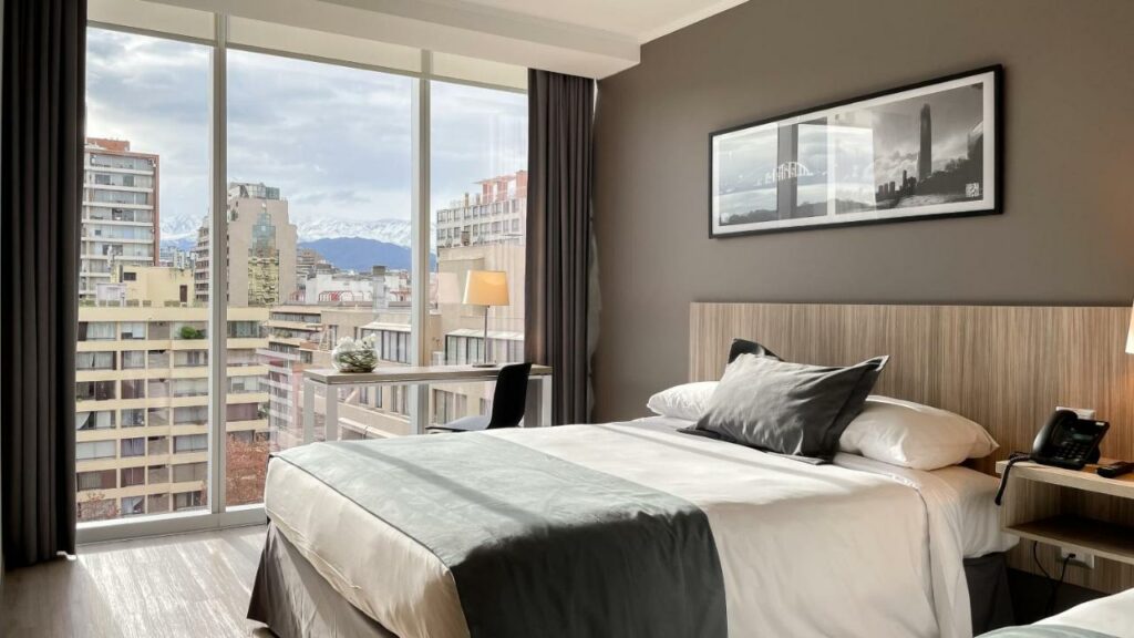 Quarto do Hotel Capital Bellet com cama de casal do lado direito da imagem e do lado direito da cama uma mesa com cadeira com vista para a cidade. Representa hotéis 3 estrelas em Santiago.