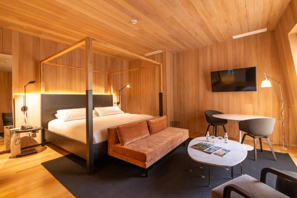 Quarto do Hotel Magnolia Santiago com cama de casal do lado esquerdo da imagem no pé da cama um sofá e do lado direto uma mesa redonda com duas cadeiras. Representa hotéis 5 estrelas em Santiago.