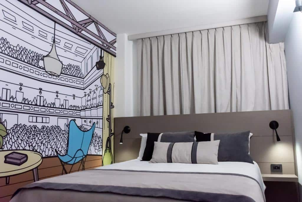 quarto do ibis Styles Buenos Aires Florida com cama de casal, mesinha e luminária de ambos os lados, janela acima e nas paredes um papel de parede com desenhos de um teatro ao estilo cartum