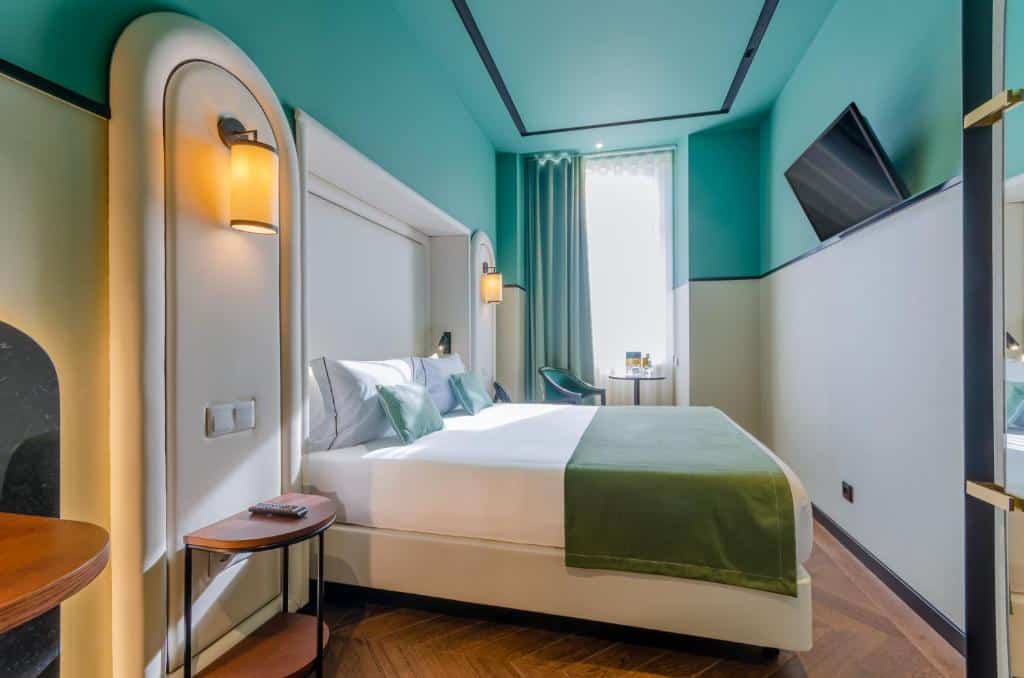 Quarto do Nicola Rossio Hotel com uma cama de casal, uma janela com cortinas e uma pequena mesinha com uma cadeira estofada, de frente com a cama há uma televisão e uma mesinha de cabeceira com uma luminária presa na parede