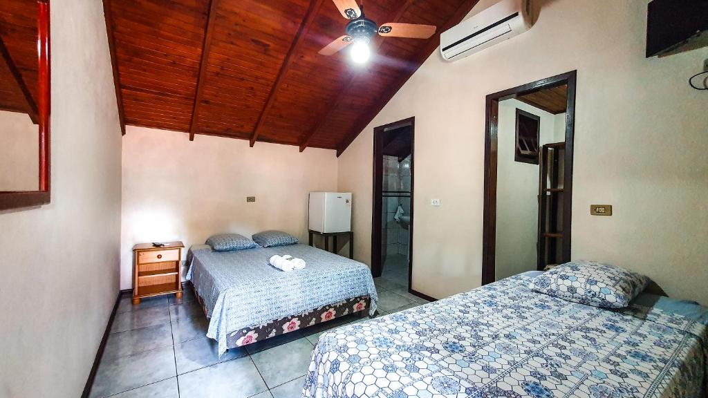 Quarto da Pousada do Alemão com duas camas de casal. A cama de casa ao fundo do quarto tem uma mesa de cabeceira do lado e um frigobar do outro. Já a cama de casal mais a frente tem uma tv por perto e um ar-condicionado. No quarto há duas portas abertas que dá acesso a outros cômodos.