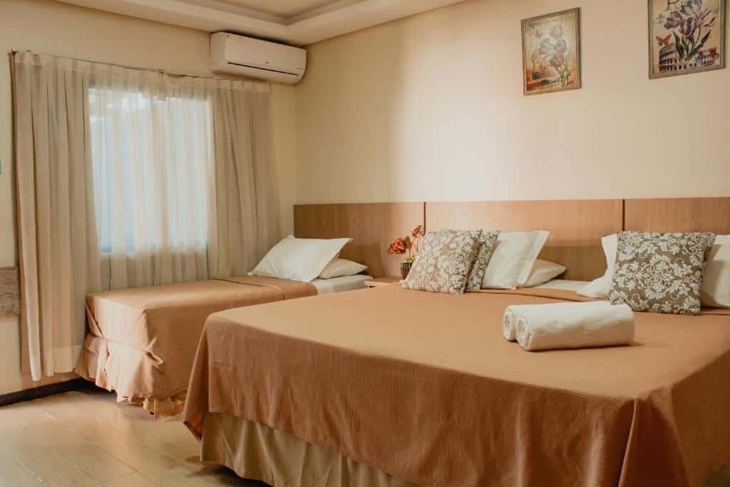Quarto da Pousada Nanai com uma cama de casal do lado direito e uma cama de solteiro do lado esquerdo do quarto. Do lado da cama de solteiro tem uma janela com cortinas e em cima um ar-condicionado.