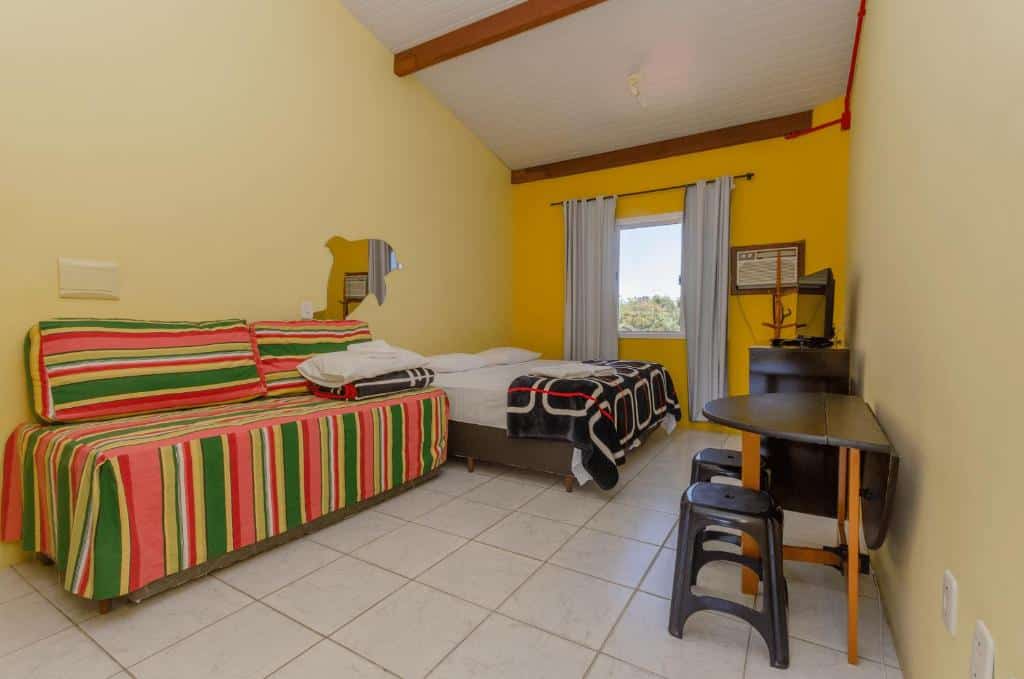Quarto da Pousada Recanto Dom Arthur com uma sala com um sofá listrado, uma cama de casal, uma escrivaninha, duas banquetas e uma janela com cortinas brancas.