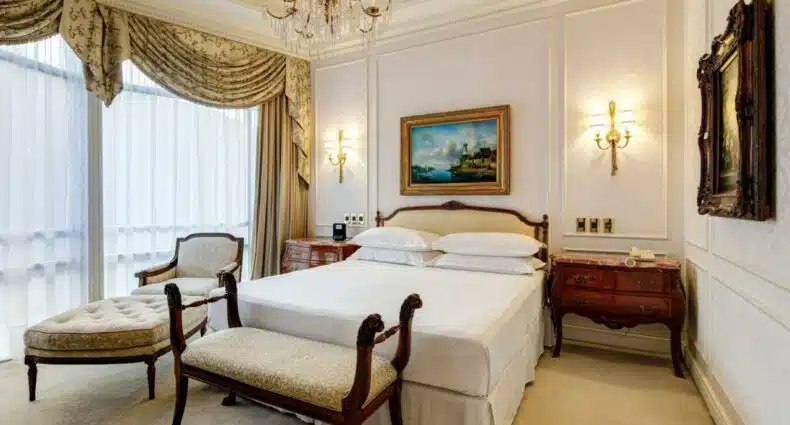 Imagem do Sheraton Santiago Hotel & Convention Center com cama de casal no centro do quarto com uma comoda em cada lado com um banco estofado do pé da cama. Representa hotéis 5 estrelas em Santiago.