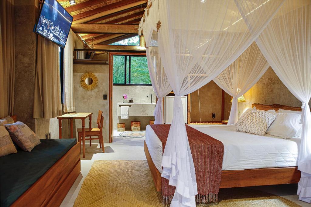 Quarto luxuoso do Villa Manakás com uma cama de casal com dossel, decoração rústica e moderna, uma televisão, duas janelas e um sofá com almofadas