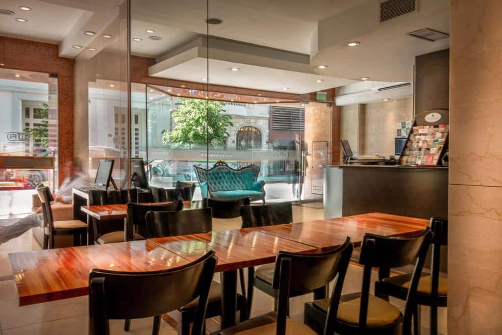 recepção e área para café da manhã com mesas e cadeiras de madeira, há um sofá em estilo clássico e os ambientes são separados por vidros no Up Viamonte Hotel, um dos hotéis 3 estrelas em Buenos Aires