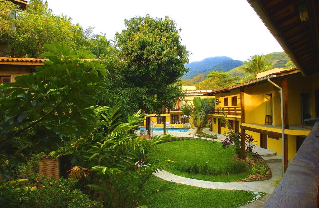 Foto exterior do Residencial Vilamar. Está mostrando um jardim com árvores e arbustos. No fundo está a estrutura da pousada, na cor amarela. Também é possível ver a piscina.