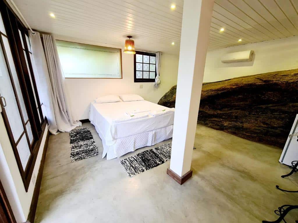 Foto do quarto em Residencial Vilamar. Ilustra o post sobre Onde ficar em Ilhabela. O ambiente é espaçoso. Há uma cama no centro, sendo box e de casal. Há uma janela do lado esquerdo do lado da cama. Um ar-condicionado está na parede à direita. Há uma coluna na frente da cama, no centro do quarto.