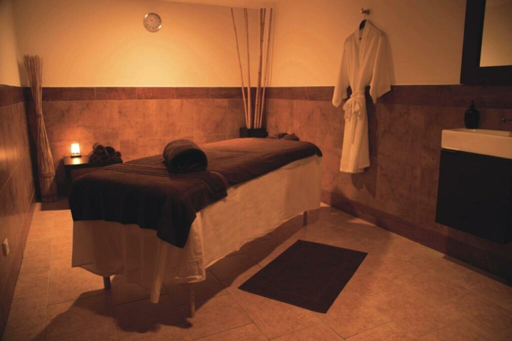 Quarto de massagem no spa do Riu Palace Aruba - All Inclusive. Uma cama preparada está bem no meio do ambiente, que também conta com um roupão pendurado na parede e uma lux mais escura e alaranjada.