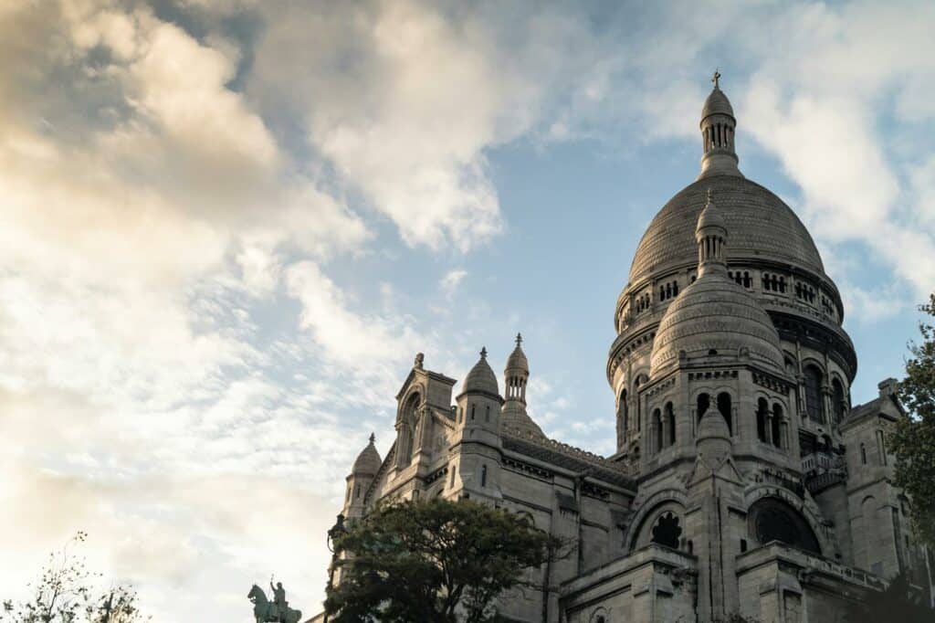 A imagem mostra uma vista parcial da Basílica de Sacré-Cœur, em Paris, destacando suas cúpulas brancas em contraste com o céu azul com nuvens espalhadas. A basílica, com sua arquitetura imponente e detalhada, é retratada de um ângulo baixo, realçando suas formas arredondadas e detalhes ornamentais. A luz suave do final do dia ilumina a cena, criando uma atmosfera serena. Ao fundo, o céu acrescenta profundidade à imagem, enquanto uma árvore aparece parcialmente no canto inferior direito.