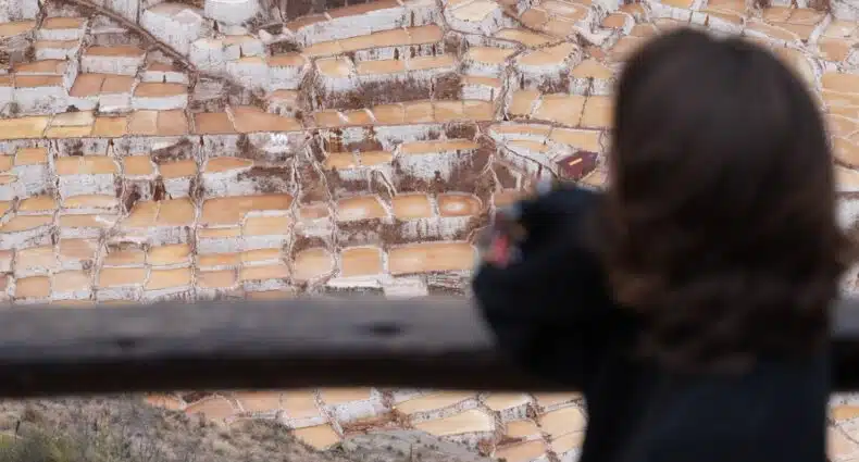 vista panorâmica de cima das salinas de maras com uma mulher de cabelo curto e repicado de costas, no canto direito da imagem, olhando para as piscinas de sal lá embaixo