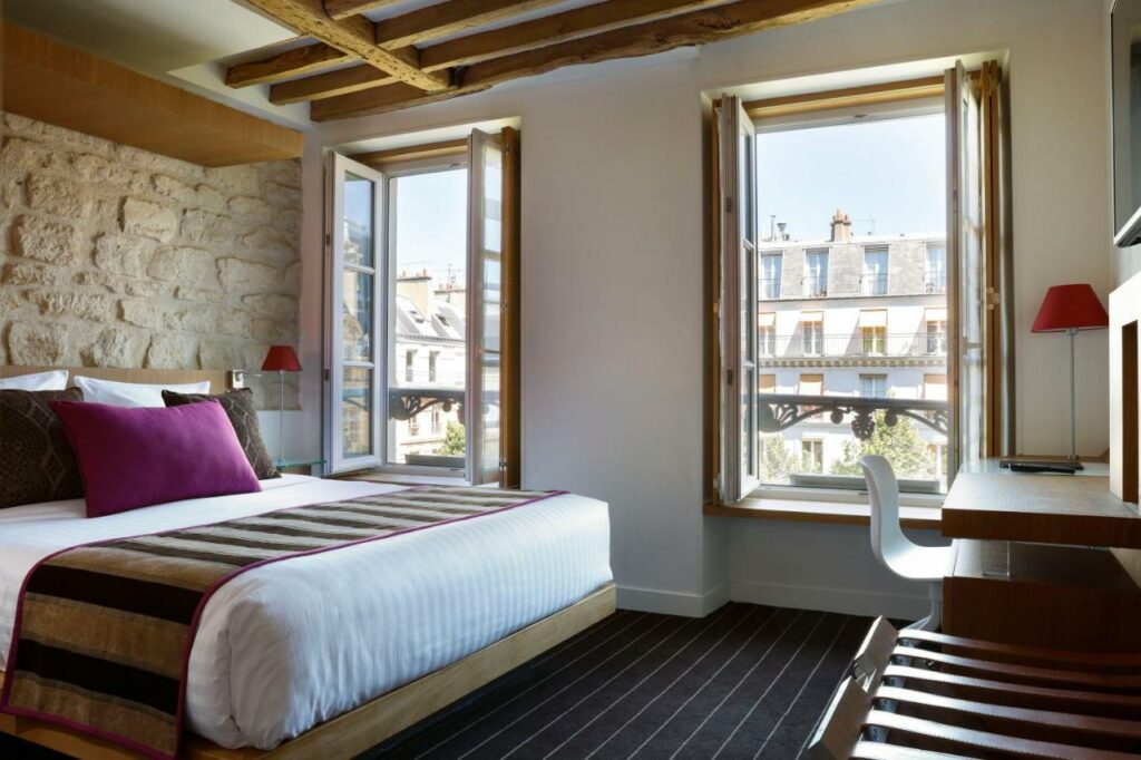 Quarto do Select Hotel, um dos hotéis 4 estrelas em Paris. Uma cama de casal está ao lado esquerdo da imagem com mesinhas de cabeceira com luminárias dos dois lados. Na lateral direita da cama ficam duas janelas com cortinas, e logo em frente à cama há uma mesa de trabalho com cadeira, abajur e TV na parede acima.