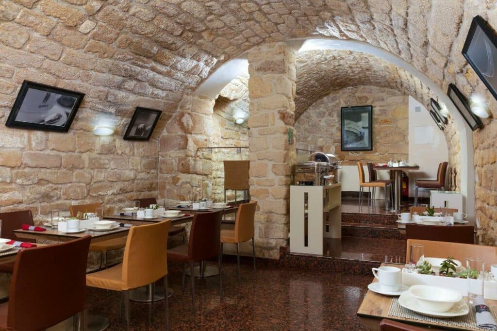 Salão de café da manhã numa antiga adega abobadada do Select Hotel. As mesas com cadeiras e sofás estão dispostas nas laterais do local, e ao fundo fica a bancada com itens do café. Há também quadrinhos nas paredes,