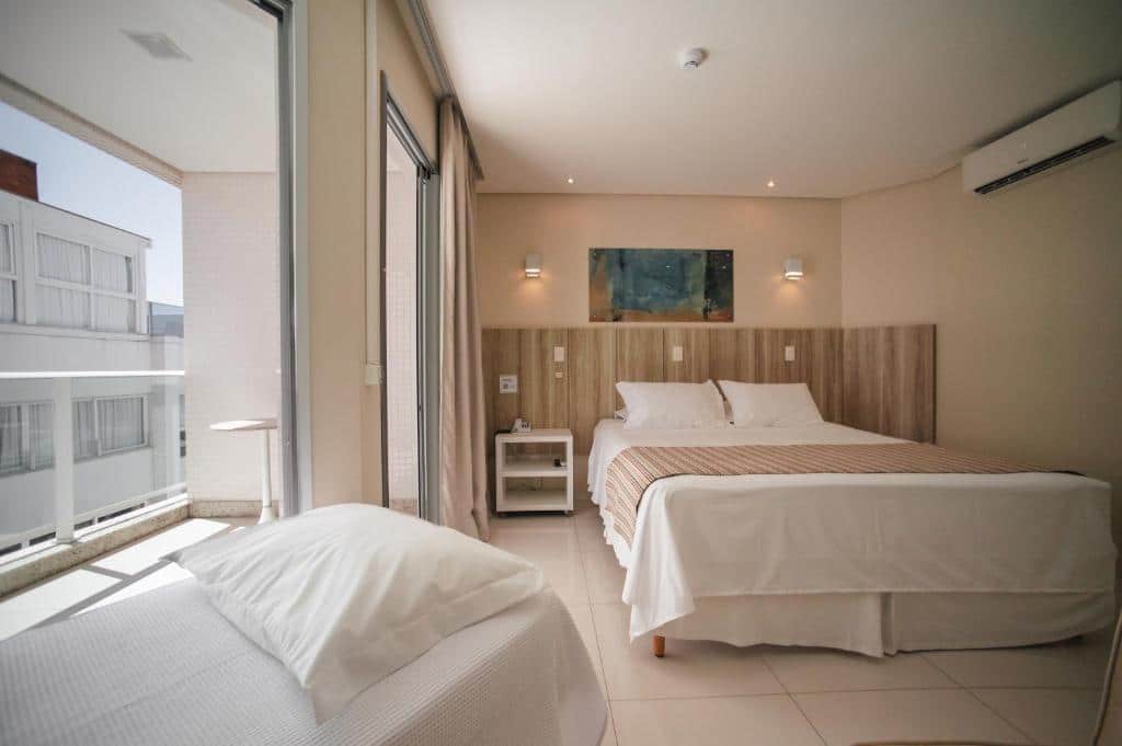 Foto do quarto do hotel Slaviero Ingleses Convention. Ilustra o post sobre Onde ficar em Florianópolis. Há uma cama box de casal no centro do quarto. Na parede atrás dela há um quadro e luminárias. Do lado direito da cama há uma porta que leva à uma varanda.