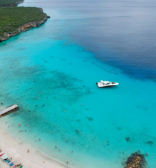 Praia Porto Mari em Curaçao, o mar é azul muito claro, sendo possível ver alguns corais, a areia é branca e há pessoas no local, para representar Ilhas ABC (Aruba, Bonaire e Curaçao)