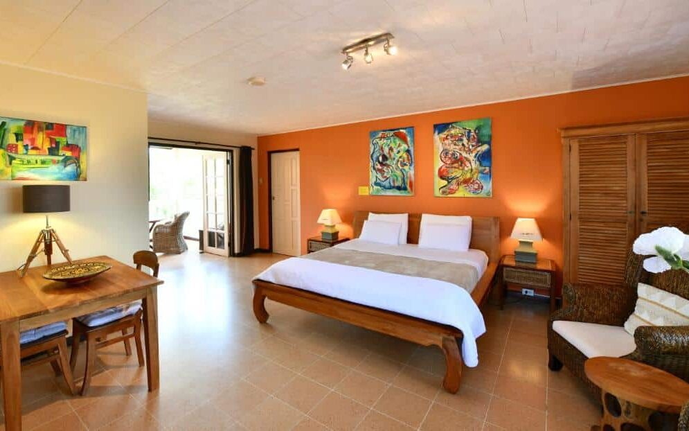 Quarto amplo do Wonders Boutique Hotel, outra opção enttre os hotpeis baratos em Aruba. Em uma parede inteira na cor laranja há dois quadros coloridos enfeitando a cabeceira da cama de casal com dois abajures, um de cada lado da cama. Há ainda um guarda-roupa de madeira no canto direito e uma mesa de madeira com duas cadeiras no canto esquerdo.