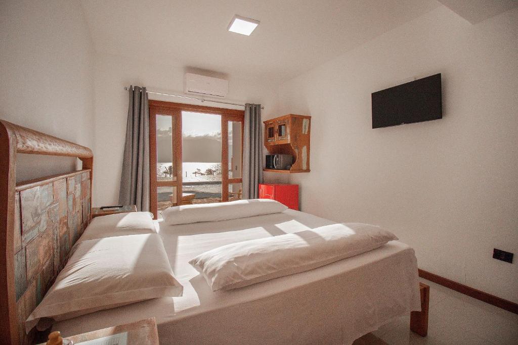 Foto do quarto em suítes vista pro mar ilhabela. Ilustra o post sobre Onde ficar em Ilhabela. Há uma cama na esquerda, box e de casal. Na frente dela há uma parede com televisão. No fundo do quarto há um frigobar e um armário com micro-ondas, além de uma porta com varanda e vista para o mar.