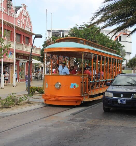Um ônibus de Aruba no meio, em Oranjestade. Ao lado direito um carro, ao lado esquerdo a calçada e um prédio antigo. Foto para ilustrar post sobre transportes em Aruba.