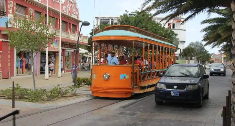 Um ônibus de Aruba no meio, em Oranjestade. Ao lado direito um carro, ao lado esquerdo a calçada e um prédio antigo. Foto para ilustrar post sobre transportes em Aruba.