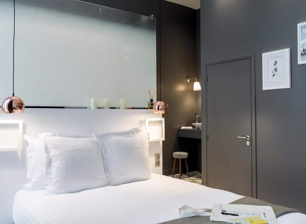 Quarto do 1er Etage Opéra, um dos hotéis perto da Galeria Lafayette. Uma cama de casal está encostada na parede ao lado esquerdo com mesinhas de cabeceira e luminárias dos dois lados. Ao lado direito fica a parede com a porta, e ao fundo é possível ver a pia do banheiro.