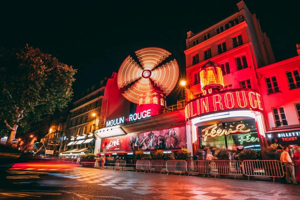 Fachada do Moulin Rouge iluminada em vermelho à noite. Várias grades protegem a fila do local, e há letreiros e um moinho de vento se movimentando ali. Essa é uma das opções de o que fazer em Paris.