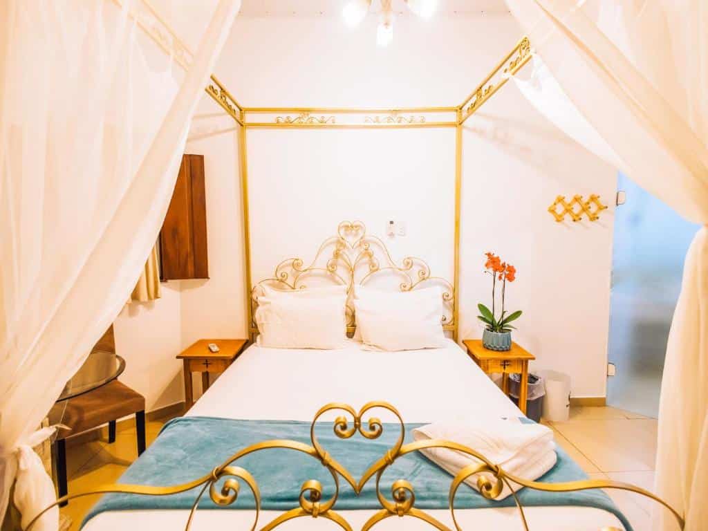 Foto do quarto em VELINN Hotel Santa Tereza. Ilustra o post sobre Onde ficar em Ilhabela. Há uma cama box de casal no centro da foto. Os detalhes da cama são em estilo clássico. Dos dois lados da cama há uma mesa e cabeceira de madeira, e a da direita há um vaso com flores.