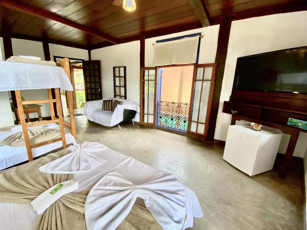 Foto do quarto em Vila Pequeá Ilhabela. Ilustra o post sobre Onde ficar em Ilhabela. Há uma cama box de casal e uma beliche no canto esquerdo do quarto. Na direita há um frigobar e uma TV. Mais ao fundo há uma janela com vista e um sofá, e também a porta do quarto.