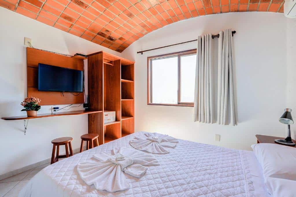 Foto do quarto do hotel Village Paraíso. Ilustra o post sobre Onde ficar em Florianópolis. No canto inferior direito há uma cama box de casal. Na frente há um painel com televisão, mesa e bancos. Do lado há um armário. Uma janela está do lado direito da cama.