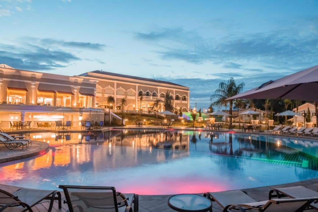 Área da piscina iluminada ao ar livre, ao redor cadeiras de tomar sol, mesas, guarda-sóis e no fundo o Wish Foz do Iguaçu.
