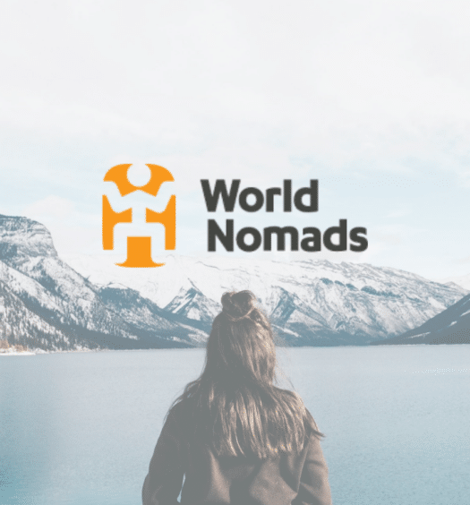 Uma mulher parada em frente a um grande lago com montanhas nevadas ao fundo, para representar seguro viagem da World Nomads