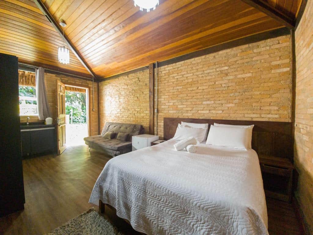 Foto do quarto em Yannai Chalé Praia. Ilustra o post sobre Onde ficar em Ilhabela. As paredes são de tijolos, dando um ar rústico. A cama é de casal e box. Do lado esquerdo dela há um frigobar e um sofá, além de uma mini cozinha.