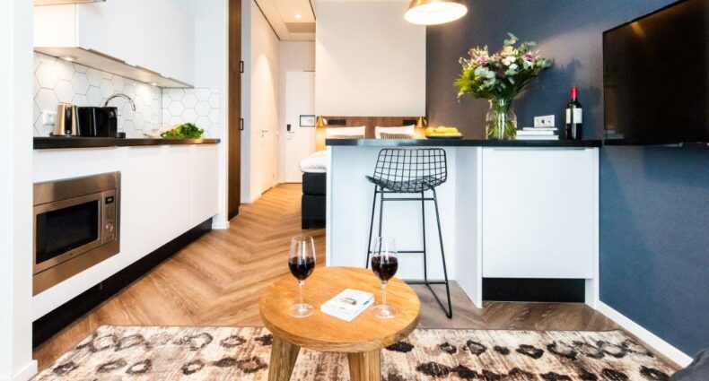 Interior de um estúdio do YAYS Docklands de Amsterdã, um dos aluguel de temporada, com uma mesinha de centro com duas taças de vinho em cima. Atrás há um balcão com uma banqueta na frente, uma TV suspensa, a cama mais ao fundo e, à esquerda, tem a cozinha com pia, forno e armários.