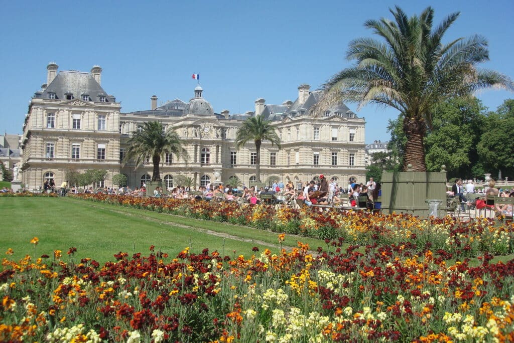 Flores coloridas ao redor de um gramado com um palácio de fundo. Várias pessoas se reunem perto das flores para tirar fotos, e há mais árvores espalhadas pelo local. Ao fundo há também uma bandeira da França hasteada no palácio. - Foto: Adriano Ribeiro via Flickr