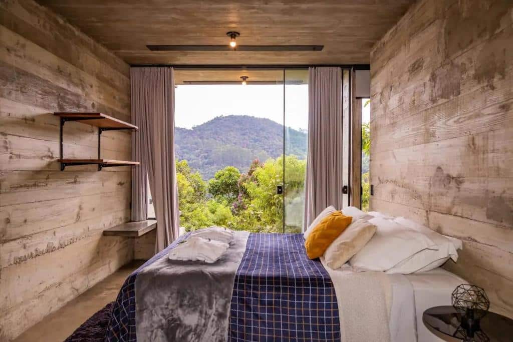 Quarto do chalé Alto de Santa Barbara. Uma cama de casal está ao lado da porta de uma varanda com vista para as montanhas. Na parede em frente a cama estão duas prateleiras. 