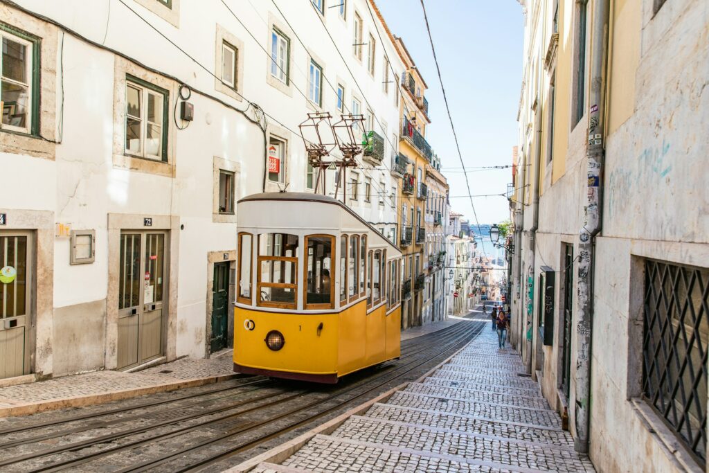 Um bonde amarelo característico de Lisboa subindo uma ladeira, na mesma rua, mais ao fundo, é possível ver o mar e as construções de época