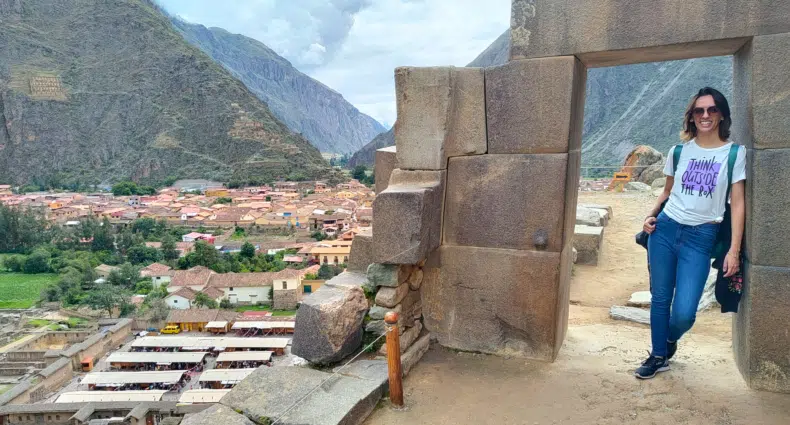 vista panorâmica da cidade de ollantaytambo no vale sagrado dos incas. No lado direito da imagem há um arco de pedra em formato de porta, onde uma mulher está apoiada com os ombros, posando e sorrindo para a câmera. No lado direito da imagem é possível enxergar a cidade lá embaixo