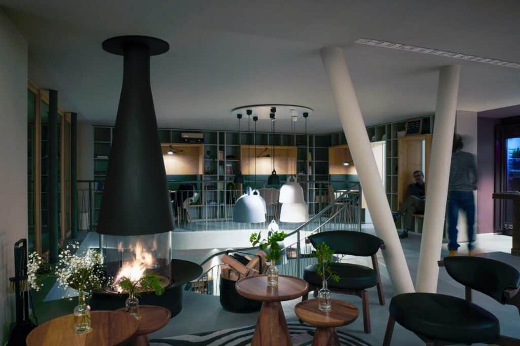 Lounge compartilhado do Carina – Design&Lifestyle hotel com mesas e cadeiras no ambiente e do lado esquerdo uma lareira.