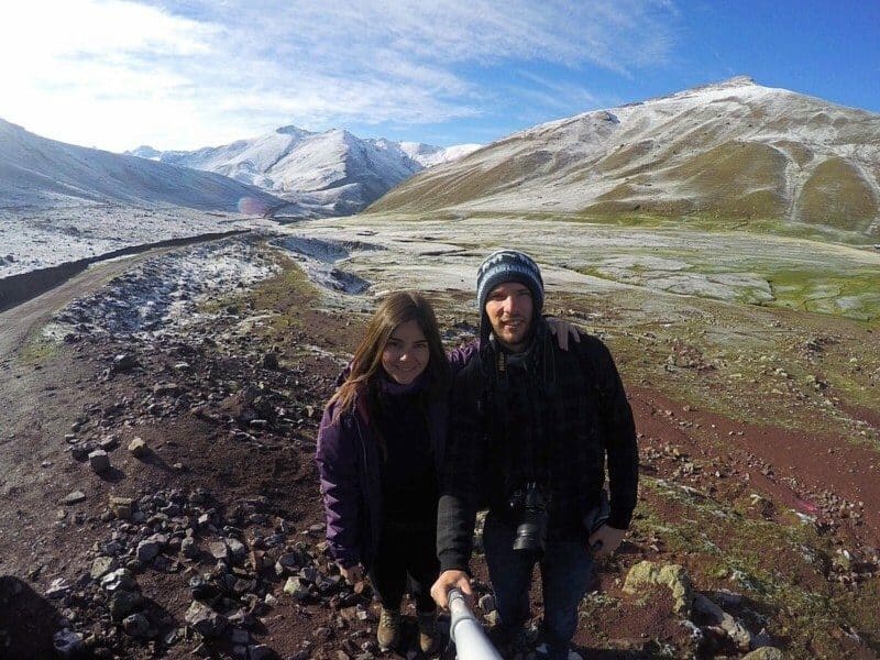Casal, sorrindo, com as montanhas de Ausangate atrás, durante o dia. A mulher está com um dos braços no ombro do homem, e o homem está segurando com uma das mãos um extensor de câmera. Representa o que fazer no Peru