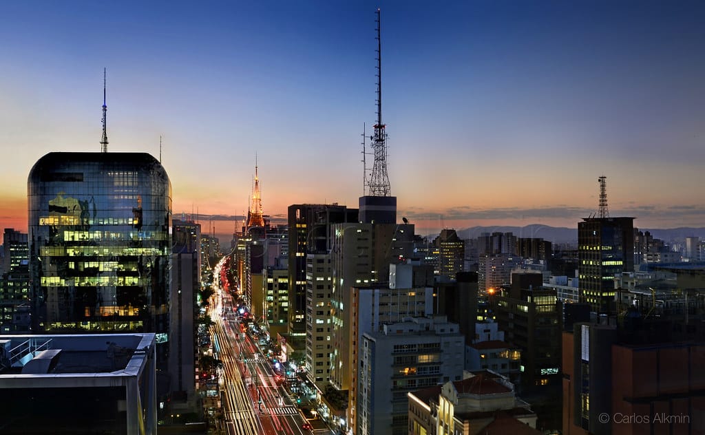 Imagem de cima da Avenida Paulista no entardecer. A avenida iluminada com o movimento dos carros e ao redor os prédios com janelas iluminadas.