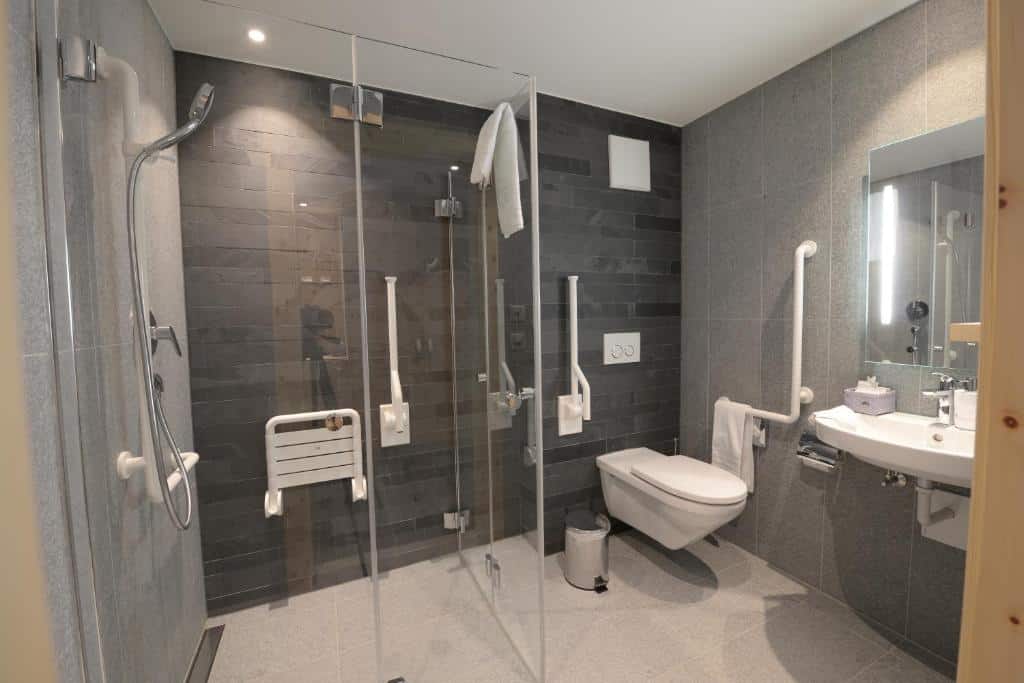 Banheiro com acessibilidade do Alpenhotel Fleurs de Zermatt com vaso sanitário com barras de apoio do lado direito da imagem do lado esquerdo o chuveiro com barras de apoio. Representa hotéis em Zermatt.