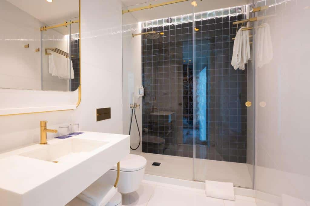 Banheiro do Blue Liberdade Hotel com pia branca, box de vidro, vaso sanitário branco e tudo com detalhes em dourado, há também um amplo espelho e toalhas brancas disponíveis, para representar hotéis 3 estrelas em Lisboa