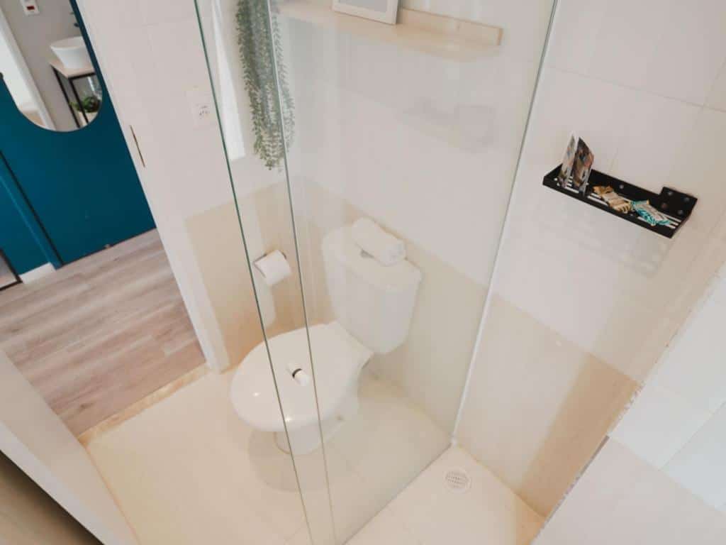 Um banheiro pequeno do Estudio do Apê Paulista, com uma porta de vidro para o chuveiro à esquerda e um vaso sanitário branco à direita. O banheiro é revestido com azulejos brancos e possui um suporte para produtos de higiene pessoal dentro do box do chuveiro. O piso do banheiro é de azulejos de tom claro, e parece haver uma passagem para outra área mais além.